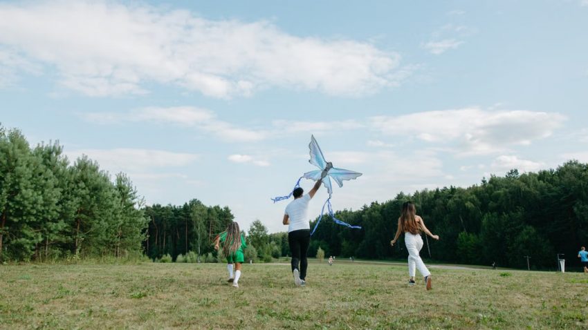 A family runs joyfully while flying a kite in a sunny park setting.